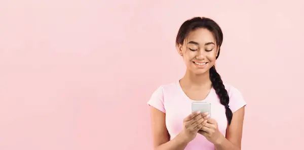 stock image A young African American girl is seen holding a cell phone up in her hand against a vibrant pink background. She appears focused on the device she is holding, copy space