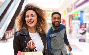 Afrika kökenli Amerikalı kadın ve erkek alışveriş merkezlerinde ellerinde alışveriş torbalarıyla dolaşıyorlar. Kadın daha fazlasını tutarken adam birkaç çanta taşıyor..