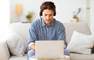 Evindeki koltukta oturan bir adam, kulaklık takıyor ve dizüstü bilgisayarla çalışıyor..