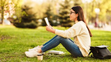 Asyalı kadın öğrenci yeşil çimlerin üzerinde oturmuş, bir kitap okumaya dalmış. Tek başına, önündeki sayfalara odaklanmış, yüzünde sakin bir ifadeyle..