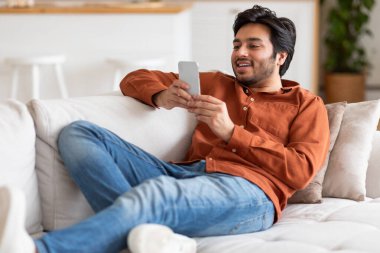 Günlük turuncu gömlek ve kot pantolon giyen genç bir adam akıllı telefonuna bakarken beyaz bir kanepeye rahatça oturur. Mutlu görünüyor ve cihazla meşgul.
