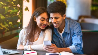 Masada oturan genç siyah bir çift, ikisi de cep telefonu ekranına odaklanmış. Cihazla meşgul görünüyorlar, muhtemelen bir şeyler okuyor ya da izliyorlar.