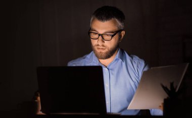 Gözlüklü ve mavi gömlekli bir adam loş bir ofiste gece geç saatlere kadar çalışıyor. Dizüstü bilgisayarına ve elinde tuttuğu bir belgeye odaklanıyor..