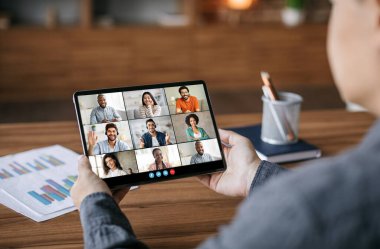 Bir kişi masada oturuyor ve bir tablet bilgisayara bakıyor. Tablette sekiz kişiyle bir video konferansı var. İnsanlar gülümsüyor ve kameraya bakıyor..
