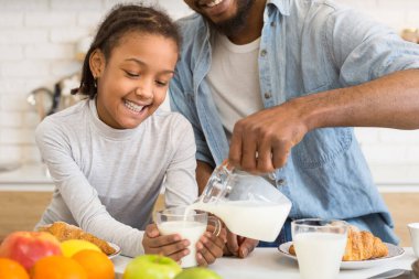 Gülümseyen bir Afrikalı Amerikalı genç kız babasının aydınlık bir mutfakta bardağa süt dökmesini izliyor.