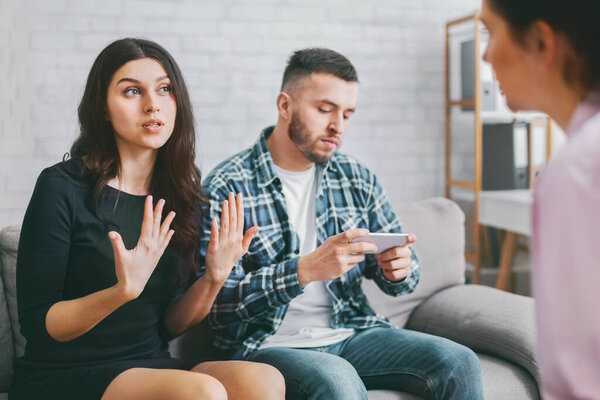 Молодая пара сидит на диване в кабинете психотерапевта. Женщина разговаривает с терапевтом, в то время как мужчина смотрит на его телефон. Женщина явно расстроена..