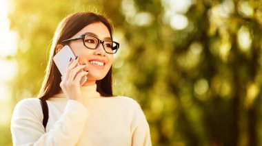 Gözlüklü Asyalı kadın cep telefonuyla konuşuyor. Cihaza konuşurken odaklanmış ve özenli görünüyor. Arkaplan tarafsız bir ortam gösteriyor