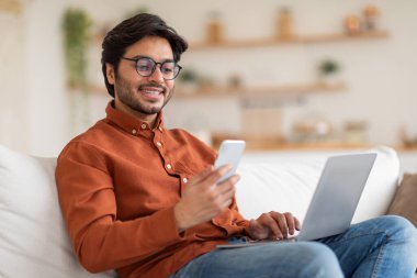 Kahverengi gömlek ve kot giyen bir adam kapalı bir yerde beyaz bir kanepede oturuyor. O, akıllı telefonunu sol eliyle ve laptopunu sağ eliyle kullanıyor. Adam gülümsüyor ve mutlu görünüyor..