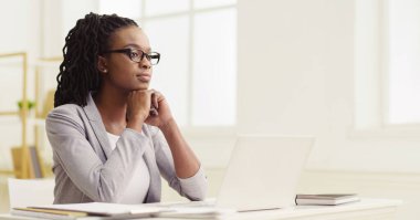 Düşünceli siyah kadın çalışan modern bir ofiste bilgisayarını kullanıyor. Keyifli ve üretken, düzgün bir masası ve parlak bir alanı var, çalışmalarından keyif aldığını gösteriyor..