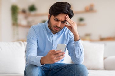 Endişeli bir ifadesi olan genç bir adam akıllı telefonunu kontrol ederken bir kanepede oturur. Mavi düğmeli gömlek ve kot pantolon giyiyor ve bir eli alnında.