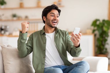 Yeşil gömlek ve kot giyen genç bir adam oturma odasındaki beyaz bir kanepede oturuyor. Akıllı telefonunu sağ elinde tutuyor ve sol yumruğunu havaya kaldırarak kutluyor.