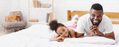 Bu resim, mutlu bir baba ve kızı yatak odasında birlikte yatarken gösteriyor. Baba karnının üstünde, doğrudan kameraya bakıyor, geniş bir gülümsemeyle, boşluğu kopyalıyor.