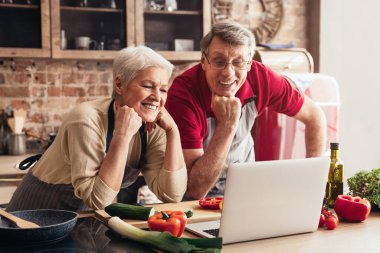 Üst sınıflardan bir çift, mutfakta yemek hazırlarken dizüstü bilgisayarda görüntülü sohbet yapıyor. Tezgahta taze sebzeler ve bir tava var..