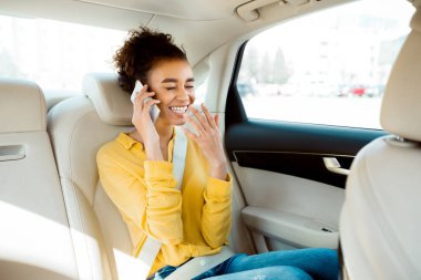 Genç siyah bir kadın arabanın arka koltuğunda oturuyor, sarı bir bluz ve kot pantolon giyiyor, cep telefonuyla konuşurken gülümsüyor. Araba bembeyaz, içi bej renginde..