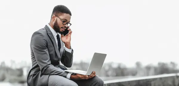 stock image Black businessman in a suit talking on a phone while working on a laptop outdoors in a professional setting.