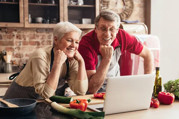 stock image A senior couple is video chatting on a laptop while preparing a meal in their kitchen. There are fresh vegetables and a pan on the countertop.