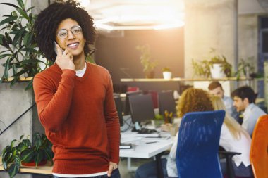 Gözlüklü neşeli bir genç adam ön planda duruyor, bir eliyle telefon ediyor, diğer eliyle de ofis geçmişiyle meşgul.