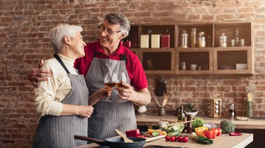 Her ikisi de önlük takan yaşlı bir çift mutfaklarında durmuş, gülümsüyor ve şarap kadehleriyle kadeh kaldırıyorlar. Mutfakta tuğla duvarlar ve ahşap dolaplar var..