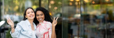 İki neşeli çoklu etnik kız arkadaş birlikte poz veriyor, kamerada barış jestleri gösteriyor, şehirde yürürken eğlenen mutlu genç kadınlar,