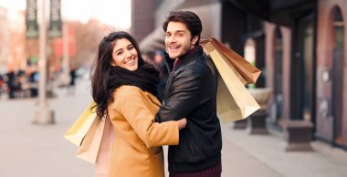 Bir adam ve bir kadın kalabalık bir caddede yürüyor, her biri alışveriş torbalarıyla dolu..