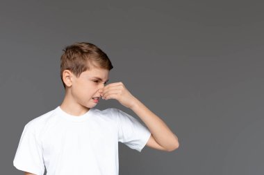 Beyaz t-shirt giyen genç bir çocuk burnunu sıkıyor. Gri bir arka planda izole edilmiş nahoş bir kokuya tepki verirken yüzünde bir iğrenme ifadesi var.