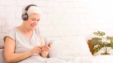 Beyaz saçlı, gülümseyen yaşlı bir kadın yatağında uzanıyor, kulaklık takıyor ve akıllı telefon kullanıyor.