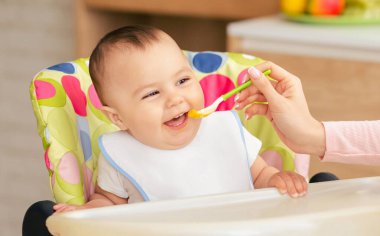 Mutlu bir bebek renkli bir sandalyede oturur ve bir yetişkin tarafından kaşıkla beslenir. Sahne aydınlık bir ev mutfağında geçiyor. Keyifli ve keyifli bir yemek zamanı öneriliyor..