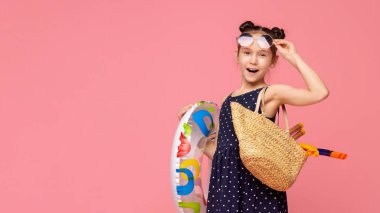 Saçları iki kalça şeklinde olan genç bir kız renkli bir yüzme yüzüğü ve pipet bir çanta tutuyor. Eğlence dolu bir yaz gününe hazır.