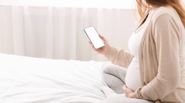 Hamile kadın yatakta oturuyor, akıllı telefon kullanarak doğumdan önce ve doğumdan sonra çocuk yetiştirmek için bilgi topluyor, boş alan.