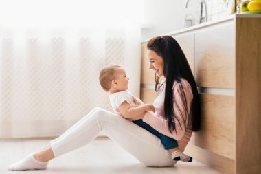 Bir kadın yerde oturuyor, şefkatle kucağında bir bebek beşiği tutuyor. Her iki birey de birbirlerine bakıyor, dokunaklı bir bağ ve bakım anı yaratıyorlar..