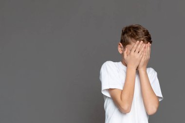 Gri bir arkaplanda, genç bir çocuk perişan ya da şaşkın bir şekilde yüzünü evrensel bir insan ifadesiyle örtüyor.