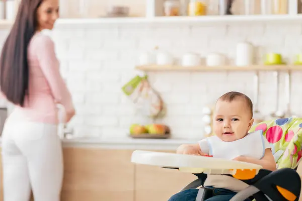 stock image A woman is standing next to a baby seated in a high chair. The woman appears to be observing or caring for the baby. The high chair is clean and modern in design, with safety straps visible.