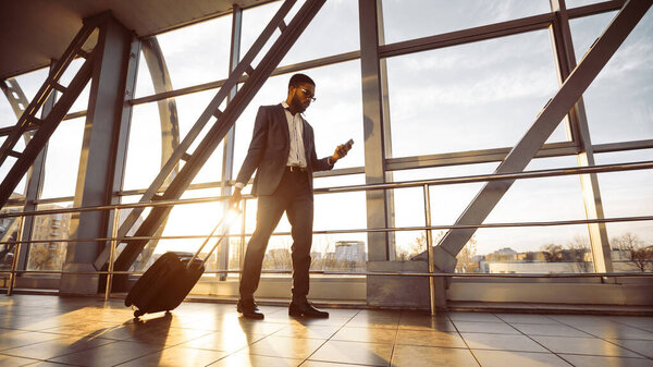 Профессиональный чёрный мужчина с багажом на телефоне в аэропорту. Атмосфера динамичная и современная, отражает деловые поездки и связи, солнечные вспышки