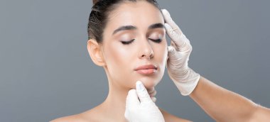 Saçları arkadan bağlanmış bir kadın beyaz eldiven giyen bir tıp uzmanı tarafından muayene ediliyor. Klinik ayarları ve profesyoneller dikkatli dokunuşlar yüz bakımına odaklanmayı öneriyor.