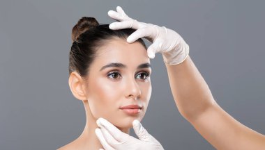 Bir dermatolog, genç bir kadının yüzünü eldivenli elleriyle nazikçe muayene eder. Klinik bir ortamda cildinde herhangi bir düzensizlik veya durum var mı diye kontrol eder..