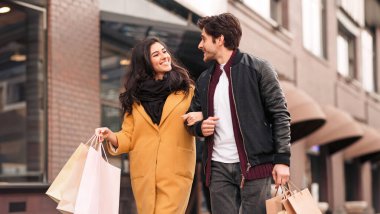 A man and woman walking down a city street while holding multiple shopping bags on a sunny day. clipart