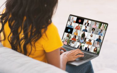 Uzun, siyah saçlı siyahi bir kadın online bir toplantıya katılırken bir kanepede oturur, ekranda diğer katılımcıların video yayınlarını gösteren bir dizüstü bilgisayar kullanır..
