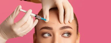 Alnına şırınga batırılan bir kadın var. Eldivenli bir el şırıngayı tutuyor ve eldivenli bir el de kadının alnını tutuyor.