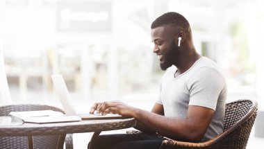 Gülümseyen bir adam bir kafedeki masada otururken dizüstü bilgisayarında çalışır. Kulaklık takıyor ve yüzünde odaklanmış bir ifade var..