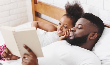 Afrikalı Amerikalı baba yatağında yatıyor ve küçük kızına kitap okuyor. Dikkatlice dinliyor ve gülümsüyor. Oda beyaz tuğla bir duvar ve ahşap bir yatak çerçevesi ile süslenmiş..