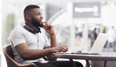 Bir adam kafeterya masasında dizüstü bilgisayarla oturuyor, düşünceli bir şekilde yan tarafa bakıyor..
