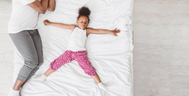 Küçük siyah bir kız babasıyla yatakta yatıyor, kolları açık şekilde sırt üstü yatıyor. Beyaz gömlek ve pembe pijama giyiyor, babası beyaz gömlek ve gri pantolon giyiyor..