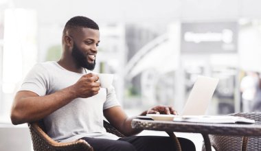 Bir adam laptopunu kullanırken gülümsüyor ve kafe masasında kahve içiyor..