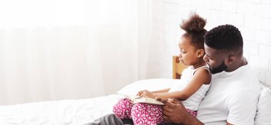 Afrika kökenli Amerikalı bir baba beyaz bir yatakta oturuyor. Kucağında oturan kızına kitap okuyor.