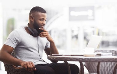 Kafe masasında oturan bir adam, kulaklıkla dizüstü bilgisayarı üzerinde çalışıyor. Düşünceli bir şekilde ekrana bakıyor, sanki çevrimiçi bir toplantı ya da projeyle ilgileniyor gibi..