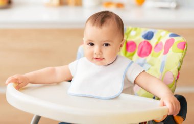 Beyaz önlük giyen sevimli bir bebek iyi ışıklandırılmış bir mutfakta yüksek bir sandalyede rahatça oturur. Bebek, sandalyenin kenarına tutunurken meraklı ve hoşnut görünüyor..