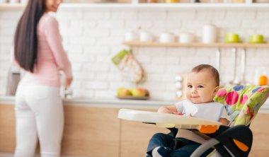 Gülümseyen bir bebek modern bir mutfakta renkli bir sandalyede rahatça otururken anne arka planda yemek hazırlamakla meşgul..