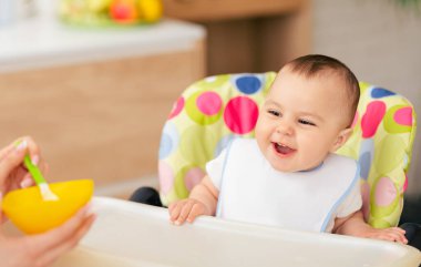 Bir anne, bebeğini mutfakta sandalyede besliyor. Sıcak ve sevecen bir ortam. Sevgi dolu bir mutfak ortamı var..