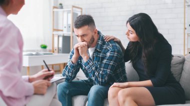 Terapi seansında koltukta oturan bir adam üzgün görünüyor ve ellerini birarada tutuyor. Bu sırada ortağı yanında oturuyor ve rahatlatıcı elini omzuna koyuyor.