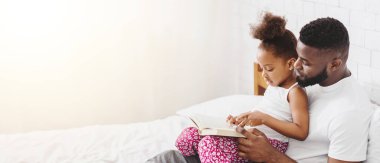 Küçük siyah bir kız babasının kucağına oturmuş yatakta kitap okuyorlar. Baba gülümsüyor ve kitabı işaret eden kızına bakıyor. Sahne sıcak ve sevgi dolu.